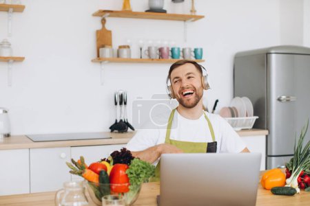 Photo for Happy man in headphones preparing salad in kitchen from fresh vegetables and looking at laptop. - Royalty Free Image