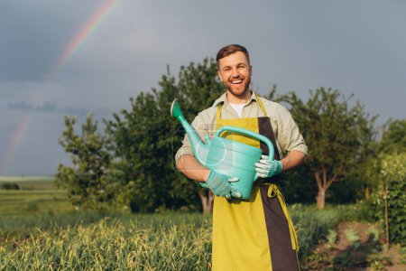 Téléchargez les photos : Heureux agriculteur masculin tenant un arrosoir dans le jardin sur fond d'arc-en-ciel d'été après la pluie - en image libre de droit