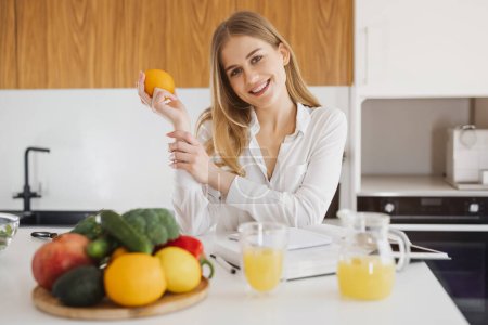 Foto de Mujer rubia linda sosteniendo naranja y mirando un libro de recetas en la cocina - Imagen libre de derechos