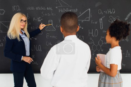 Photo for A female teacher assigns homework to school children near the blackboard. International team. Back to school. - Royalty Free Image