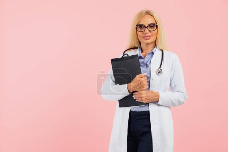 Téléchargez les photos : Femme mûre médecin dans des lunettes avec stéthoscope tenant dossier sur le fond rose avec espace de copie - en image libre de droit