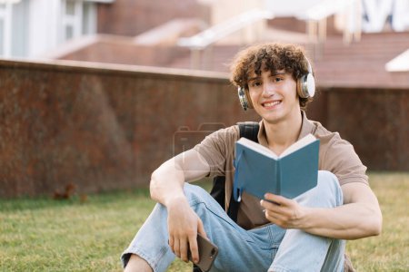 Téléchargez les photos : Attrayant étudiant masculin dans les écouteurs assis sur l'herbe faire des devoirs près de l'université le jour ensoleillé - en image libre de droit