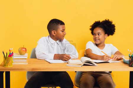 Téléchargez les photos : Heureuse écolière afro-américaine et écolier assis ensemble au bureau et étudiant sur fond jaune. Concept de retour à l'école. - en image libre de droit