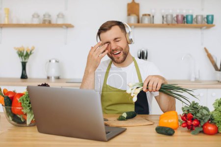 Photo for Attractive man in headphones sitting at table in kitchen preparing salad, holding onion and wipes eyes from tears. - Royalty Free Image