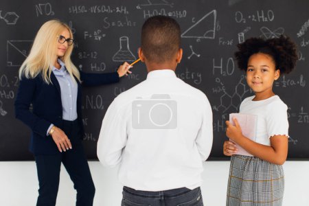 Téléchargez les photos : Une enseignante assigne des devoirs à des écoliers près du tableau noir. Équipe internationale. Retour à l'école. - en image libre de droit