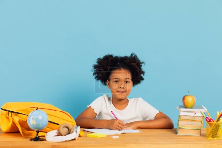 Téléchargez les photos : Heureuse écolière afro-américaine faisant ses devoirs au bureau en classe sur fond bleu. Concept de retour à l'école. - en image libre de droit