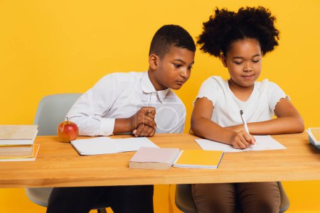 Photo for Happy african american schoolgirl and schoolboy sitting together at desk and studying on yellow background. Back to school concept. - Royalty Free Image