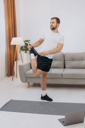 Photo for Positive athletic bearded middle-aged man stretching at home, using laptop, watching sport videos on Internet, having fitness class online, copy space. Healthy lifestyle, sport on self-isolation - Royalty Free Image
