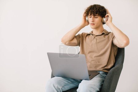 Téléchargez les photos : Jeune homme attrayant utilisant un ordinateur portable, portant des écouteurs tout en étant assis dans un fauteuil contre un mur blanc, copier l'espace. Millennial mâle communiquer en ligne, travailler ou étudier à distance sur PC portable - en image libre de droit