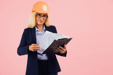 Photo for Portrait of attractive mature business woman architect or engineer in glasses use folder with documents on the pink background - Royalty Free Image
