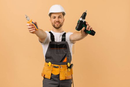 Foto de Reparador masculino positivo en casa muestra destornillador y alicates sobre fondo beige - Imagen libre de derechos