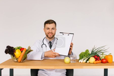 Photo for Happy doctor nutritionist sitting at workplace at desk in office among fresh vegetables and showing folder with copy space, diet plan concept - Royalty Free Image