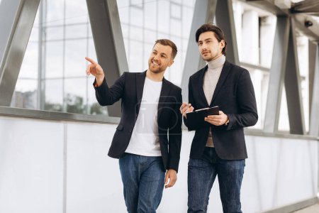 Téléchargez les photos : Deux hommes d'affaires heureux collègues se parlent tout en marchant à l'extérieur de l'immeuble de bureaux moderne. - en image libre de droit