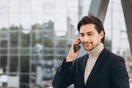 Foto de Retrato de hombre de negocios moderno guapo en traje hablando por teléfono y sonriendo en el fondo de edificios urbanos y oficinas. - Imagen libre de derechos