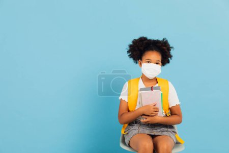 Foto de Feliz colegiala afroamericana en una máscara sentada en la silla sobre fondo azul con espacio de copia. De vuelta al concepto escolar. COVID-19 - Imagen libre de derechos