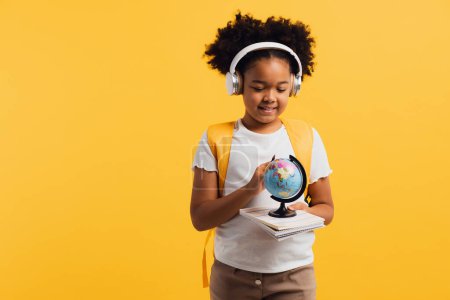 Foto de Feliz colegiala afroamericana en auriculares con mochila mirando el globo geográfico, espacio de copia. - Imagen libre de derechos