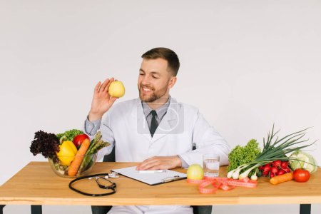 Foto de Feliz médico nutricionista sentado en la mesa de trabajo entre verduras frescas y la celebración de manzana - Imagen libre de derechos