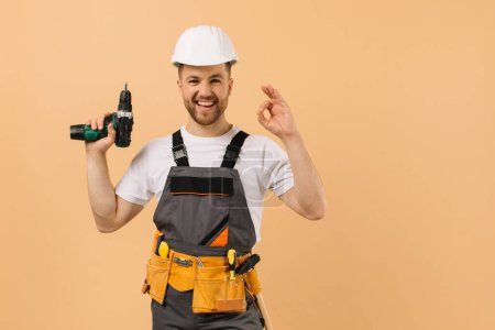 Photo for Positive male repairman at home holding a screwdriver and showing okay on a beige background - Royalty Free Image