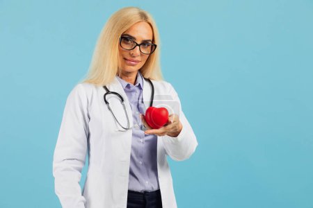 Photo for Mature woman doctor in glasses with stethoscope holding heart on the blue background with copy space - Royalty Free Image