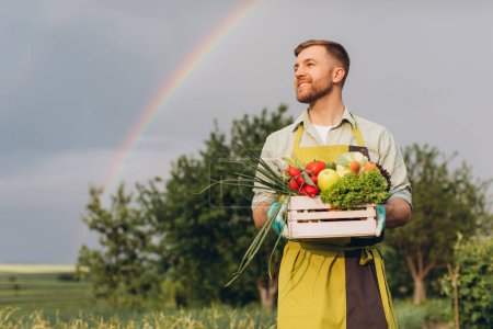 Téléchargez les photos : Heureux jardinier homme tenant panier avec des légumes frais sur fond arc-en-ciel et jardin, concept de jardinage - en image libre de droit