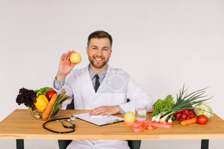 Téléchargez les photos : Heureux médecin nutritionniste assis à la table du travail parmi les légumes frais et tenant pomme - en image libre de droit