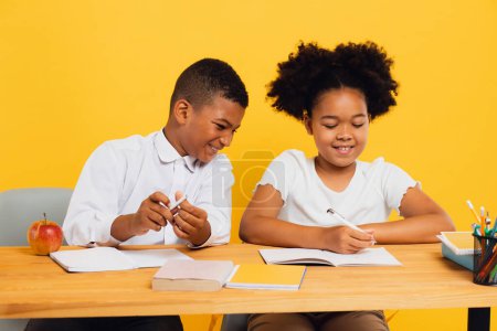 Téléchargez les photos : Joyeux écolière afro-américaine et écolier métis assis ensemble au bureau et étudiant sur fond jaune. Concept de retour à l'école. - en image libre de droit