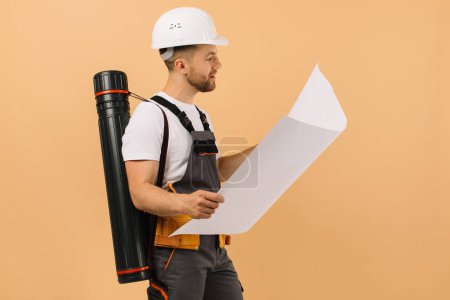 Téléchargez les photos : Ingénieur de construction positif examine un dessin et tient un tube sur un fond beige - en image libre de droit