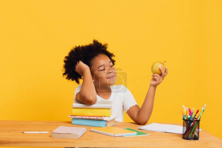 Photo for Happy African American schoolgirl doing homework and holding apple for lunch while sitting at desk. Back to school concept. - Royalty Free Image