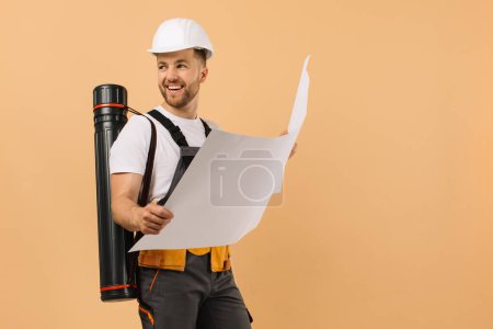 Téléchargez les photos : Ingénieur de construction positif examine un dessin et tient un tube sur un fond beige - en image libre de droit
