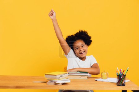 Photo for Happy African American female schoolgirl sitting at desk and stretching arm up during class. Back to school concept. - Royalty Free Image