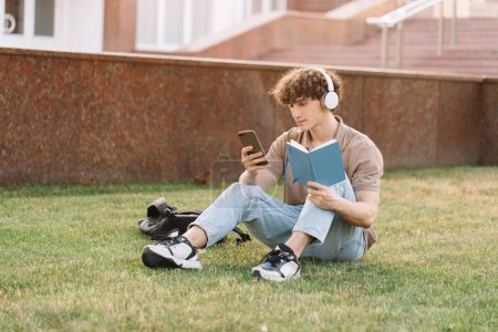 Foto de Atractivo estudiante masculino en auriculares sentados en la hierba haciendo la tarea cerca de la universidad en el día soleado - Imagen libre de derechos