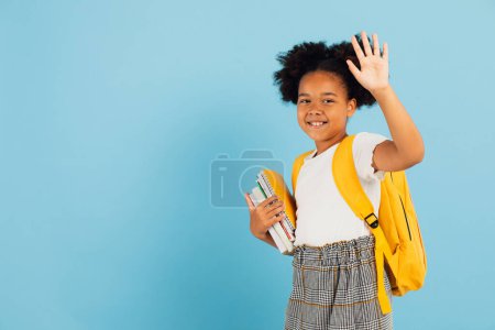 Téléchargez les photos : Bonne écolière afro-américaine fait un geste de main bienvenu sur fond bleu, concept de retour à l'école. - en image libre de droit