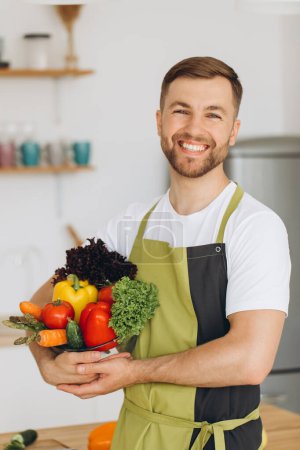 Photo for Portrait of a happy man holding a plate of fresh vegetables on the background of the kitchen at home - Royalty Free Image