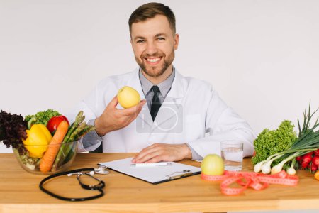 Photo for Happy doctor nutritionist sitting at workplace table among fresh vegetables and holding apple - Royalty Free Image