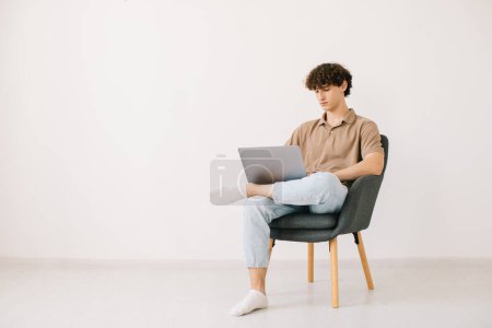 Foto de Atractivo joven que usa el ordenador portátil mientras está sentado en un sillón contra la pared blanca, copia el espacio. Hombre milenario que se comunica en línea, trabajando o estudiando remotamente en PC portátil - Imagen libre de derechos