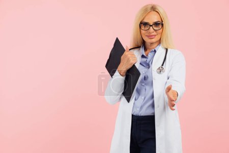 Photo for Mature woman doctor in glasses with stethoscope holding folder and give hand respect on the pink background with copy space - Royalty Free Image