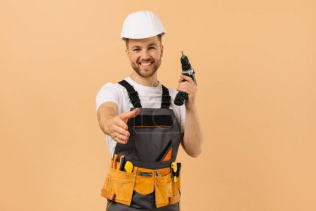 Foto de Reparador masculino positivo en casa sosteniendo un destornillador y extendiendo su mano para saludar sobre un fondo beige - Imagen libre de derechos
