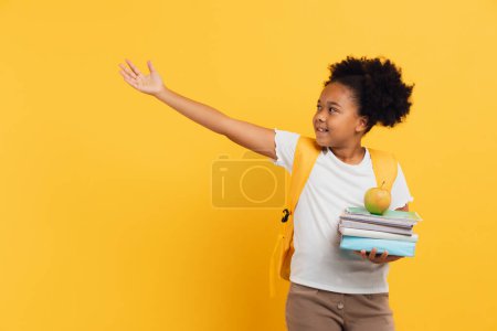 Téléchargez les photos : Écolière afro-américaine tenant des cahiers et des livres et montrant quelque chose sur fond jaune, espace de copie. - en image libre de droit