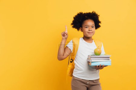 Téléchargez les photos : Écolière afro-américaine tenant des cahiers et des livres et montrant quelque chose sur fond jaune, espace de copie. - en image libre de droit