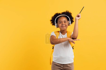 Téléchargez les photos : Heureuse écolière afro-américaine dans un casque avec sac à dos tire la main vers le haut en classe sur fond jaune, espace de copie. - en image libre de droit