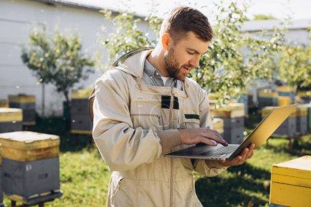 Téléchargez les photos : Homme agronome ou ingénieur en combinaison de protection travaillant sur ordinateur portable à la ferme apicole - en image libre de droit