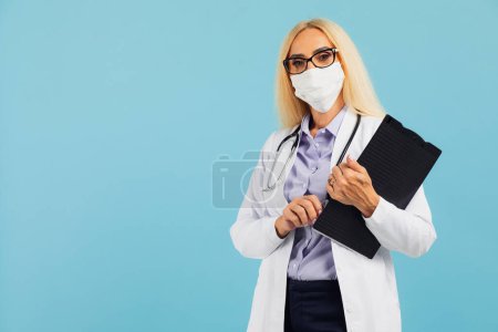 Foto de Médico mujer madura en máscara sostiene carpeta sobre fondo azul. Concepto Covid-19 - Imagen libre de derechos