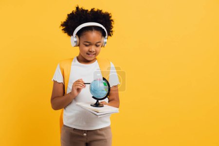 Téléchargez les photos : Heureuse écolière afro-américaine dans un casque avec sac à dos regardant le globe géographique, l'espace de copie. - en image libre de droit