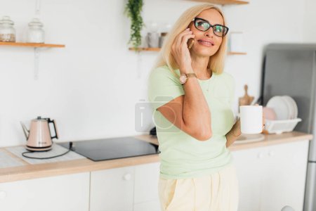 Téléchargez les photos : Moderne attrayant femme d'âge moyen parler au téléphone et boire du café dans la cuisine à la maison - en image libre de droit