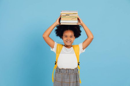 Téléchargez les photos : Happy African American écolière tenant des livres au-dessus de la tête sur fond bleu, concept de retour à l'école. - en image libre de droit