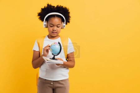 Foto de Feliz colegiala afroamericana en auriculares con mochila mirando el globo geográfico, espacio de copia. - Imagen libre de derechos