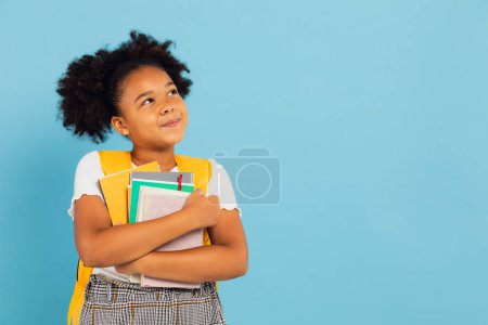 Téléchargez les photos : Heureuse écolière afro-américaine tenant des livres sur fond bleu, concept de retour à l'école. - en image libre de droit