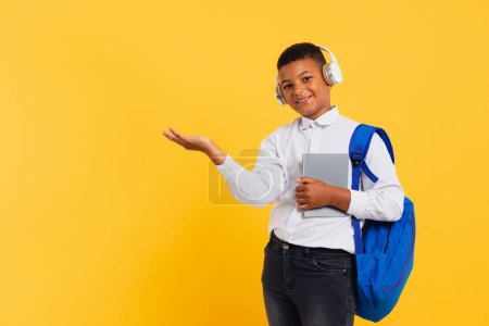 Téléchargez les photos : Heureux écolier africain portant des écouteurs et sac à dos tenant des livres et des cahiers et pointant vers l'espace de copie. Concept de retour à l'école. - en image libre de droit