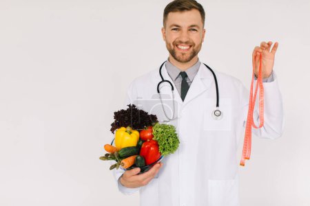 Photo for The male doctor nutritionist with stethoscope holding fresh vegetables and ruler on white background - Royalty Free Image