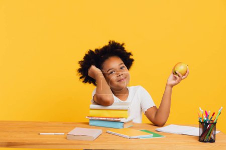 Téléchargez les photos : Heureuse écolière afro-américaine faisant ses devoirs et tenant la pomme pour le déjeuner tout en étant assise au bureau. Concept de retour à l'école. - en image libre de droit